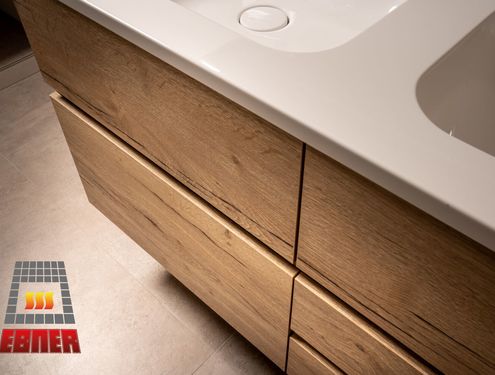 Modern, zeitlos und im Landhausstil. Durch die schlichten Fliesenfarben und den Badezimmermöbeln aus Holz wirkt das Badezimmer gemütlich und freundlich.